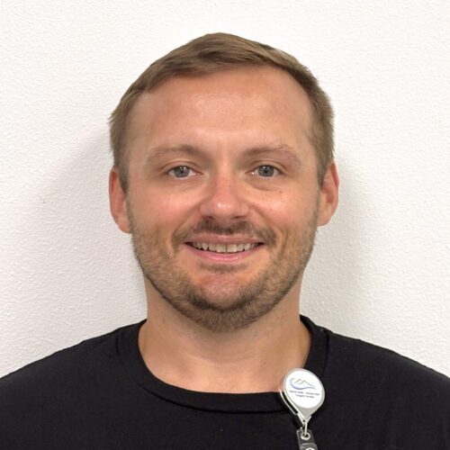 patrickB A man with short light brown hair and a trimmed beard, wearing a black shirt and a staff badge, stands against a plain white wall, smiling at the camera.